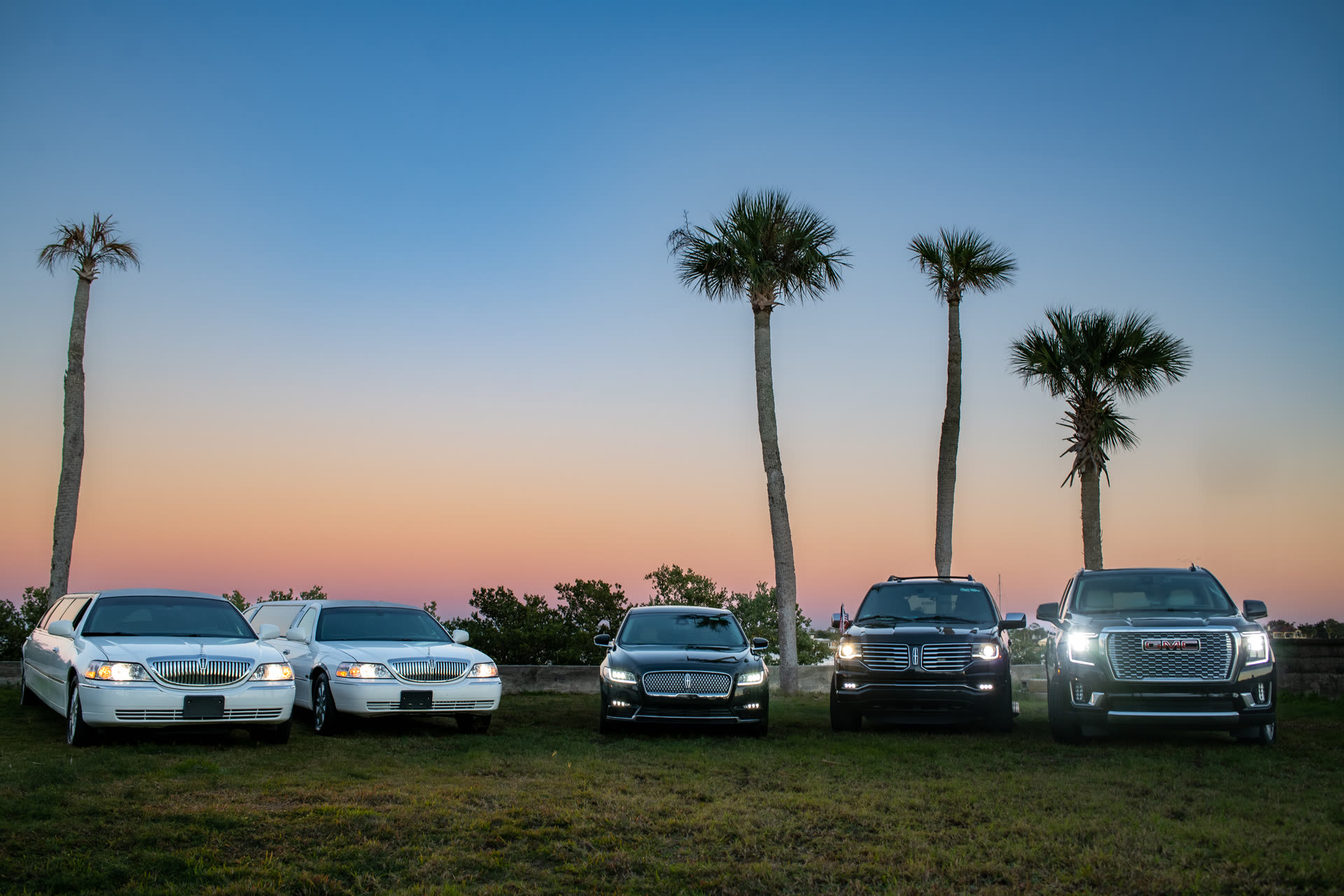The Atlantic Coach fleet — two white Lincoln stretch limousines, a black Lincoln town car, a black Lincoln Navigator SUV, and a GMC Denali — lined up at sunset with palm trees framing the scene at St. Augustine